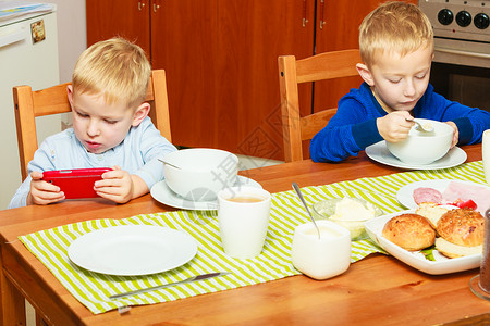 两位金发男孩子们在桌边吃早餐边玩手机图片