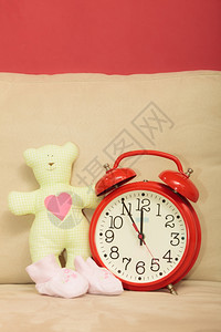 可爱的泰迪熊玩具给未出生婴儿的小鞋在家沙发上的红色闹钟图片