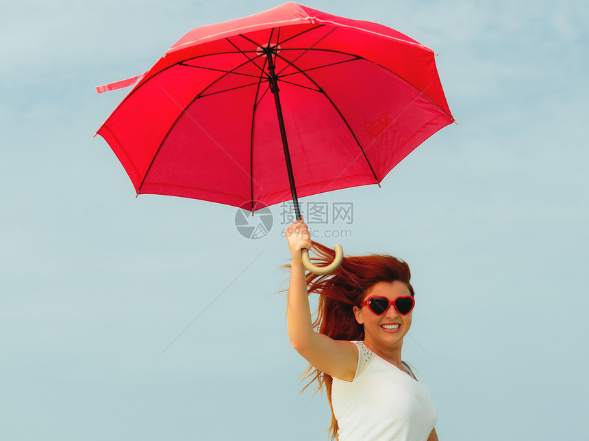 假期旅行和自由概念美丽的红发快乐女孩在海滩上拿着红伞跳跃图片