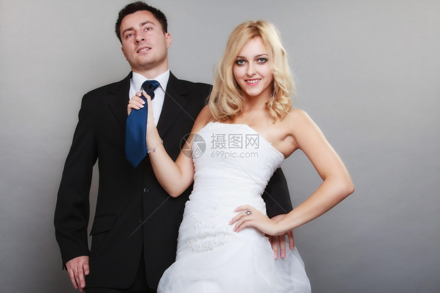 结婚日幸福的已夫妇金新娘和郎的肖像女人拉着男的领带工作室拍着灰色的背景图片