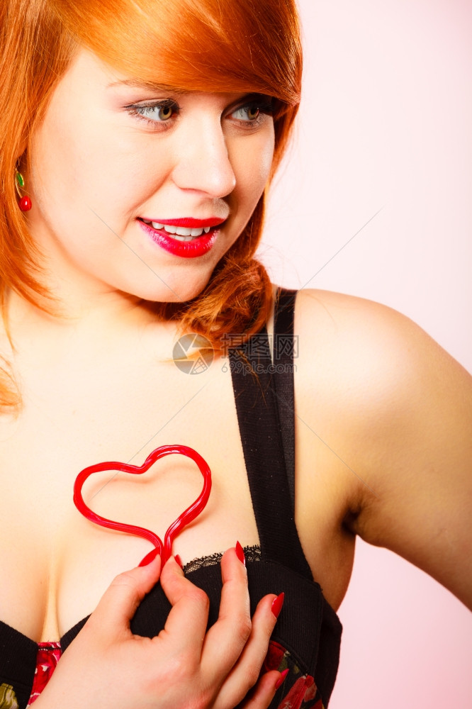 爱糖果和情人节39Day概念迷人的年轻红发女郎在工作室的粉红背景中握着糖果心图片
