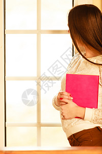 休闲文学和人的概念妇女在家里拿书时透过窗口看图片