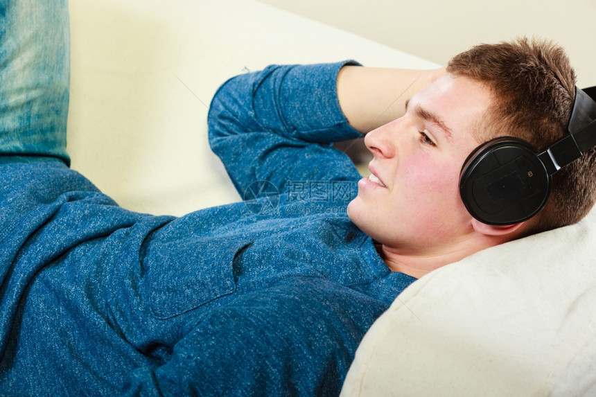 英俊的年轻人带着耳机躺在沙发上家里听音乐图片