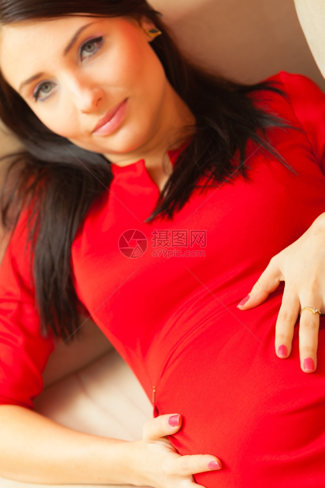 美丽的感时尚优雅的孕妇穿着红礼服在沙发上放松摸她的肚子图片