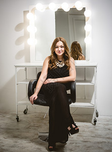 穿着长黑色裙的漂亮时髦女人坐在椅子上用灯泡对着镜子图片