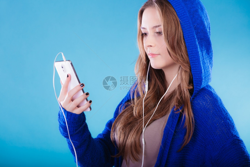 年轻女子拥有智能手机听音乐年轻时髦的长发女郎穿着兜帽放松或学习语言工作室的蓝色镜头图片