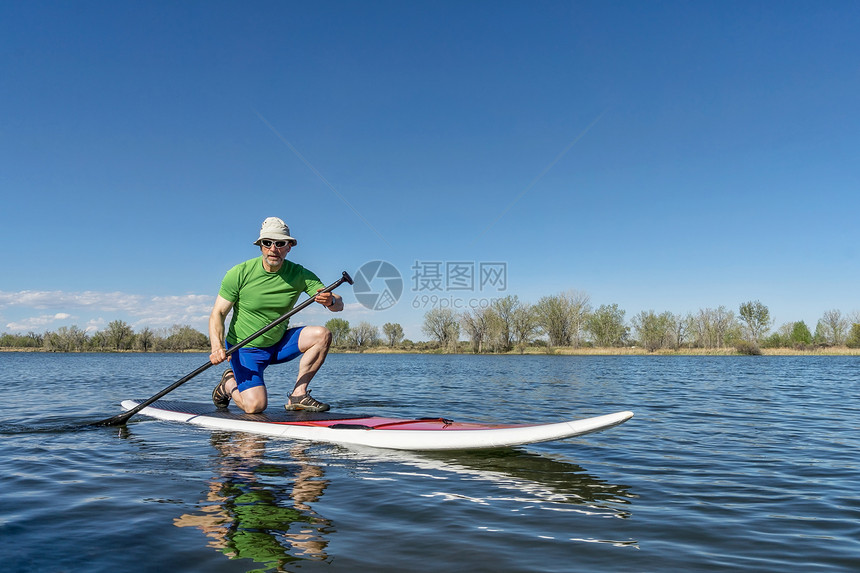 时尚的老人在平静的湖中间跪在皮划桨上划水图片