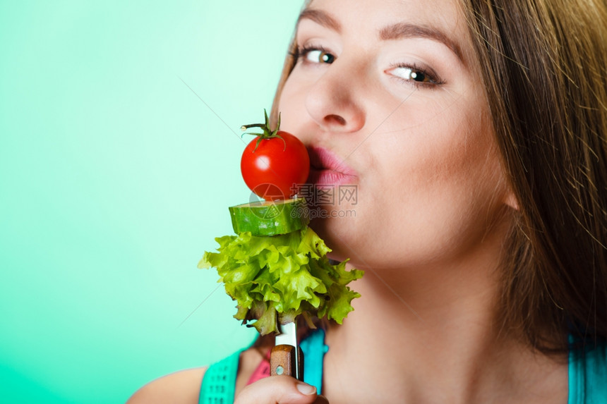 健美保和减肥概念运动健壮的女子身妇颈部有测量胶带在绿色蓝底背景上持有叉子和新鲜混合蔬菜工作室拍摄图片