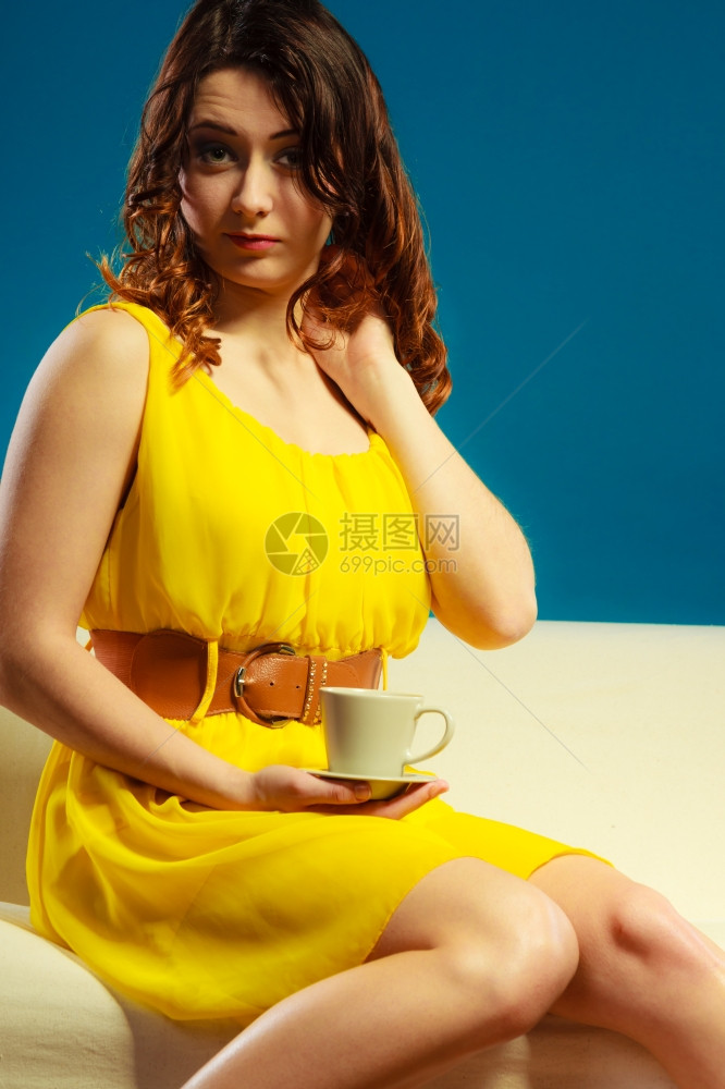 美容时装和放松概念时装女孩的黄色洋穿着热饮咖啡或茶杯坐在沙发深蓝背景的上图片