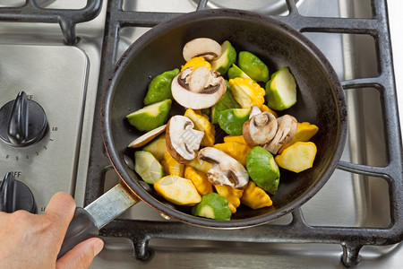 小煎锅紧握着煎锅的手专注于食物同时在炉子上用锅煮蔬菜背景