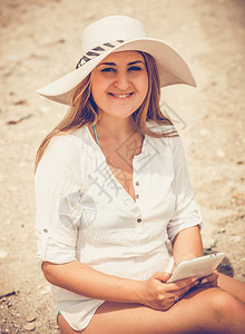 在沙滩上用平板电脑将戴着帽子的笑女人近距离剪贴肖像图片
