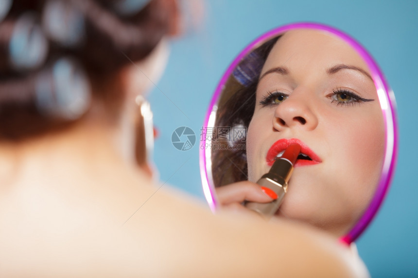 年轻女子准备参加聚会出去玩女孩用卷头发用红口涂看着镜子反光风格的蓝色背景图片