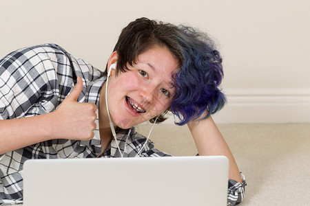 微笑的少女向前看伸出拇指举起同时使用计算机和听家里的音乐图片
