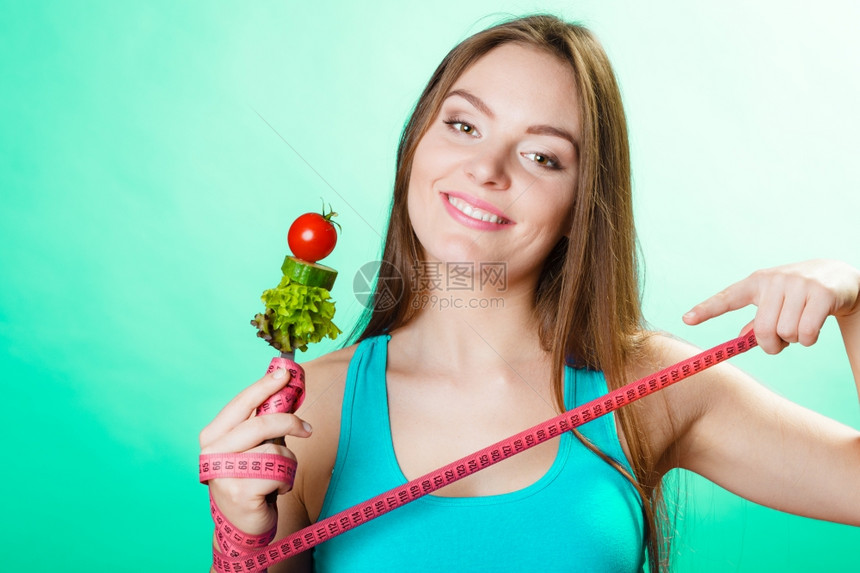 健美的身女拿着新鲜混合蔬菜的叉子和绿色蓝背景的测量胶带图片