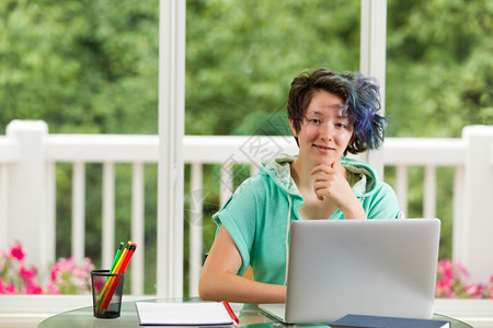 微笑的少女向前看电脑书本和铅笔放在前台深处的大窗户绿树和鲜花模糊不清图片