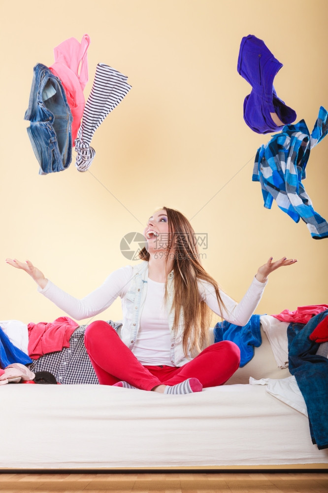 坐在沙发上客厅里扔衣服的快乐女人被许多衣服包围的年轻女孩家里的混乱和七八糟图片