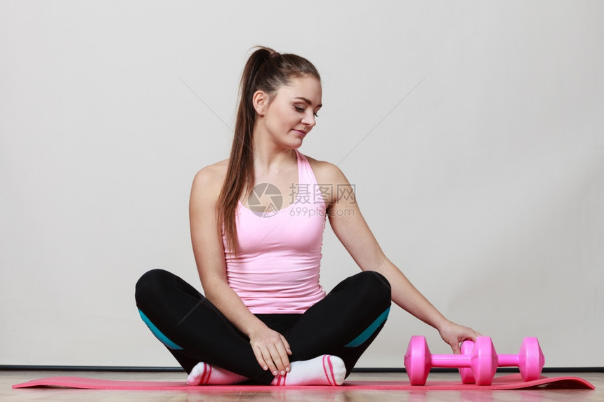 适合身体健康的女孩坐在有哑铃的运动垫子上与哑铃一起运动训练灰色背景图片