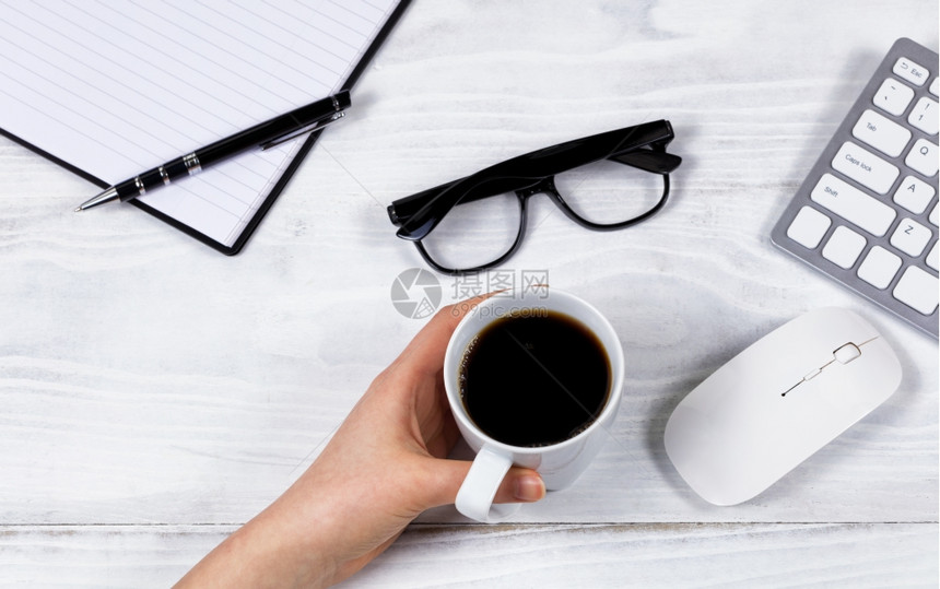 在白色桌面上用电脑键盘鼠标阅读眼镜笔记本和在白色桌面上举着黑咖啡杯的女手头视线图片