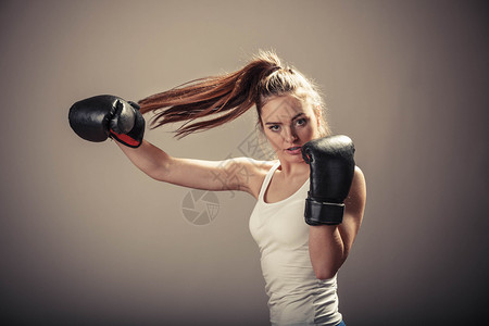 精力充沛的年轻女子用拳击手套打架与对手拳击运动和体力建设强健的年轻女子打敌有马尾的女子戴白箱顶和拳击手套背景图片