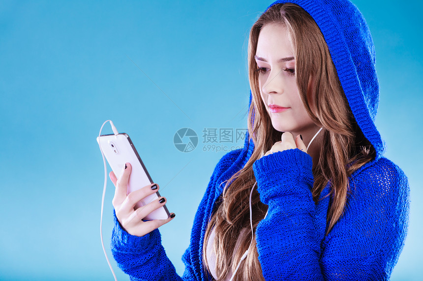 年轻女子有智能手机听音乐年轻时髦的长发女郎戴兜帽放松或学习语言工作室拍蓝色图片