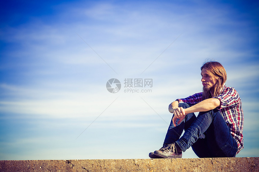 长头发在户外天空背景下放松的人长头发穿着格衬衫的人在阳光明媚的风天与蓝对立时坐在水泥墙上的在户外放松图片