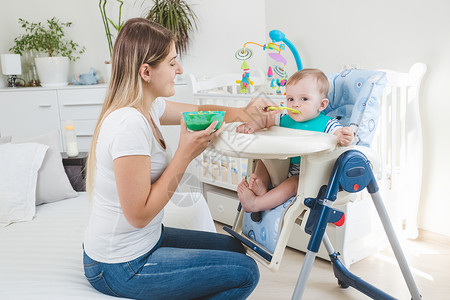 美丽的年轻母亲用粥喂她的婴儿男孩坐在高椅子上用粥煮图片