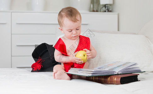 穿着毕业帽子和丝带的有趣婴儿拿着苹果看大书图片