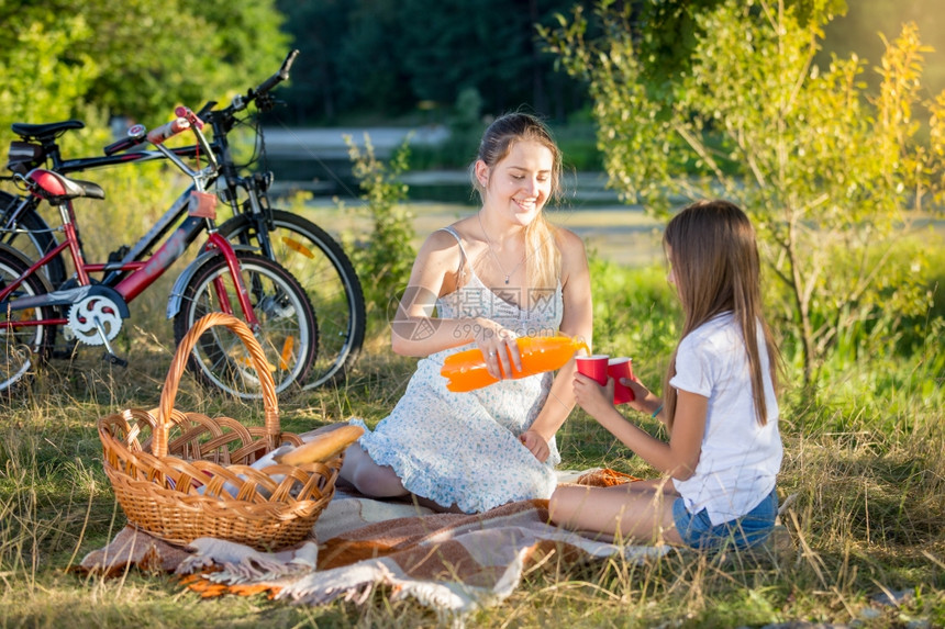 和女儿一起在河边野餐的年轻快乐母亲在女儿的杯子里倒橙汁图片