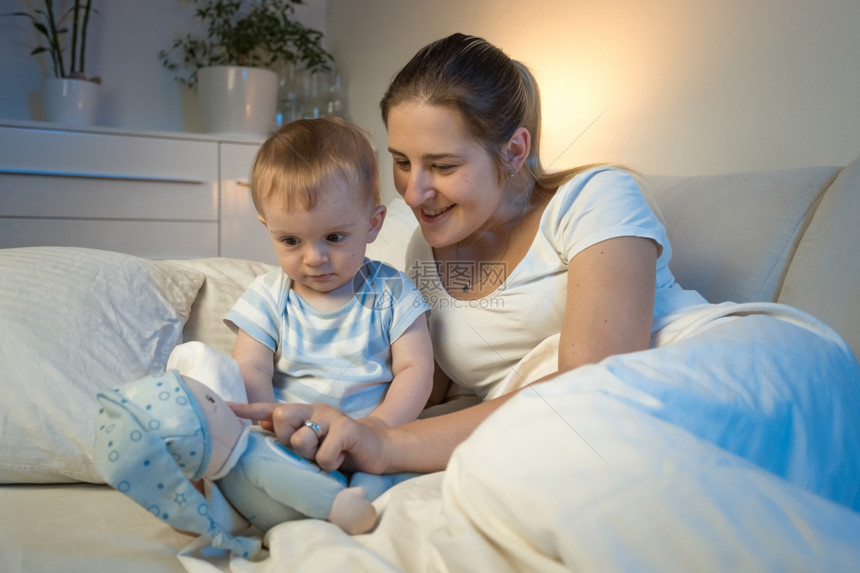 快乐笑的母亲与婴儿一起玩耍晚上躺在床玩具图片