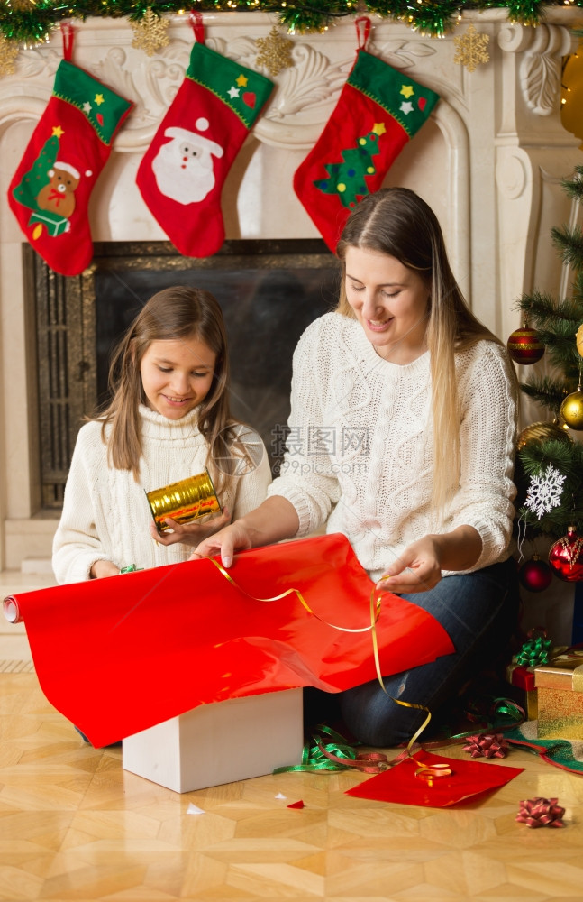 美丽的笑女孩和母亲将圣诞礼物包装在红纸和金丝带上图片
