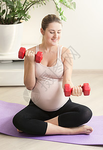 快乐的孕妇在家健身席上用红色重量在健身垫上运动图片