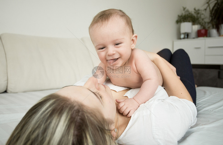 年轻微笑的母亲肖像躺在床上和她6个月的儿子玩耍图片