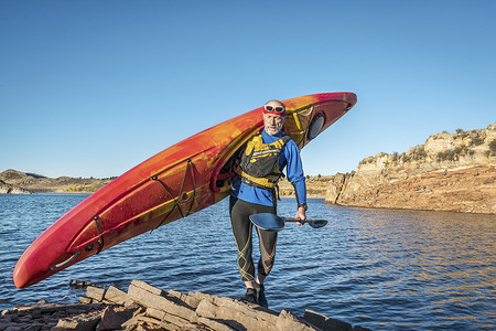 老年男在岩石湖岸上背着一条多彩的河水皮艇图片