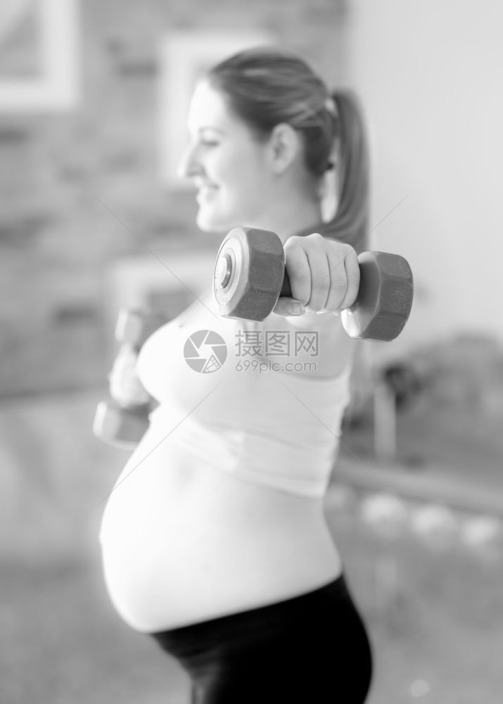 孕妇在健身房与哑铃一起锻炼时微笑着的孕妇黑色和白形象图片