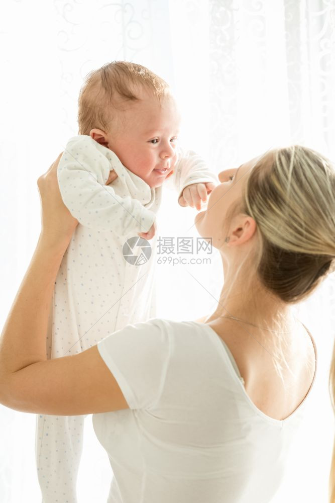 快乐的年轻母亲抱着她三个月大的婴儿在窗边图片