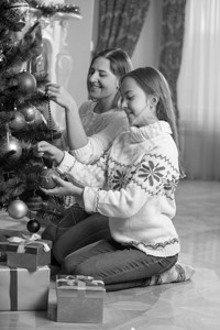年轻快乐的母亲和女儿坐在起居室地板上并装饰圣诞树的黑白画面图片