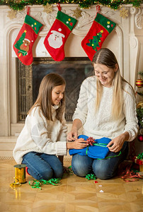 快乐的母亲和女儿坐在壁炉的地板上为圣诞礼物打包毛衣图片