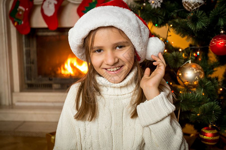 穿着圣诞老人帽子的可爱女孩肖像假扮在客厅燃烧的壁炉上图片