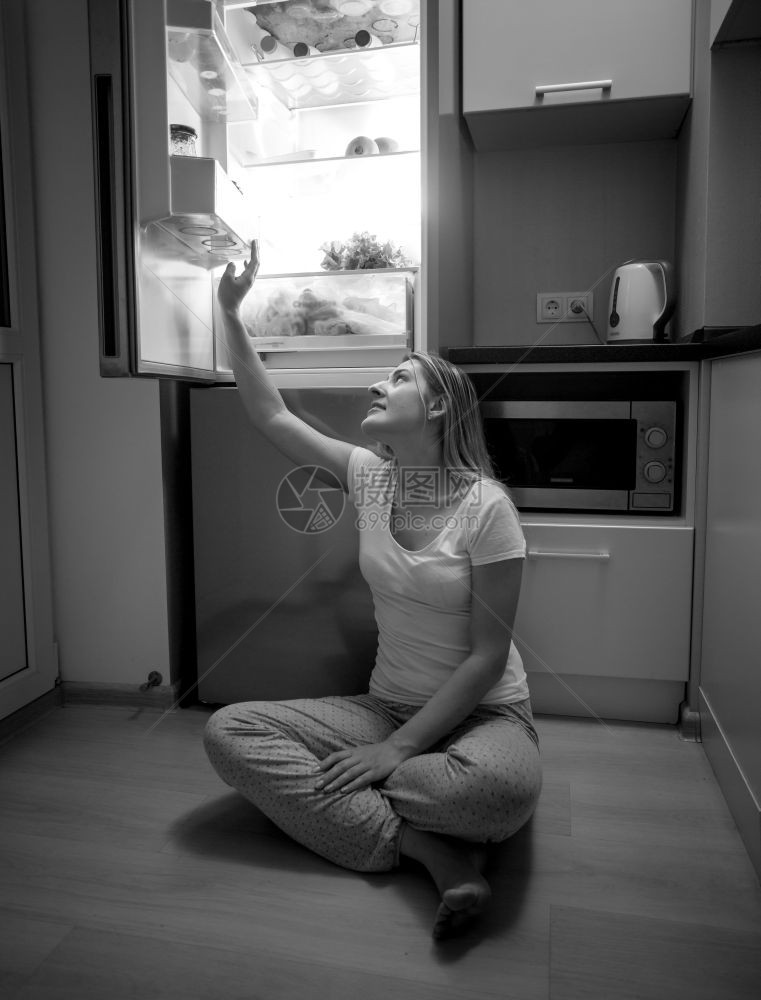 深夜坐在地板上从露天冰箱中取食物的年轻妇女黑白画面图片