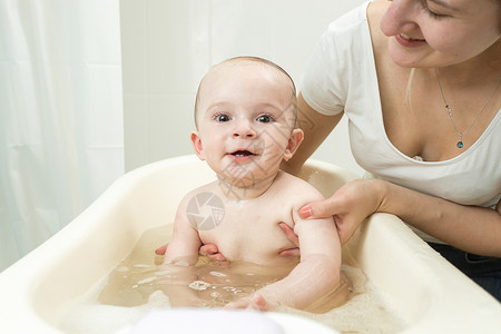 年轻微笑的母亲在塑料浴池中洗她可爱的婴儿图片