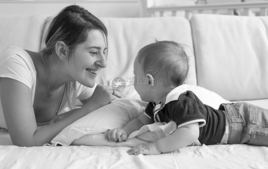 快乐的年轻母亲单色肖像躺在床上看着对方与婴儿男孩躺在床上图片