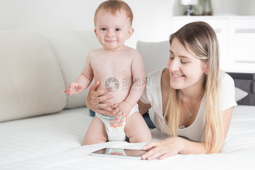 使用数字平板电脑在床上微笑的母亲和婴儿男孩图片