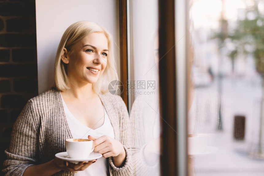 站在窗边的年轻女人拿着一杯咖啡看外面笑自然的户外灯光站在窗边的女士拿着一杯咖啡图片