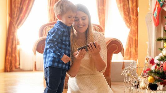 快乐的年轻母亲在圣诞树旁边和她的小儿子一起观看智能手机视频图片