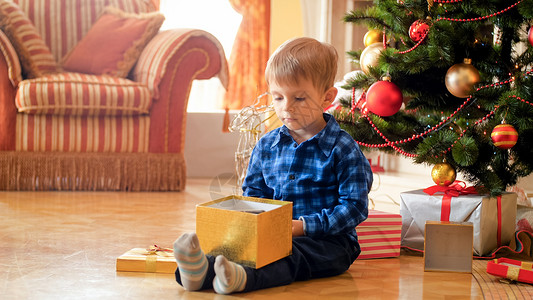 坐在圣诞树旁边的托德勒男孩看着圣诞老人的礼物盒里面图片