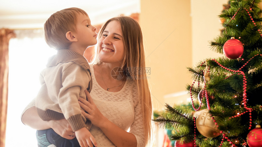 欢笑的母亲在客厅圣诞树上抱着她的小儿子图片