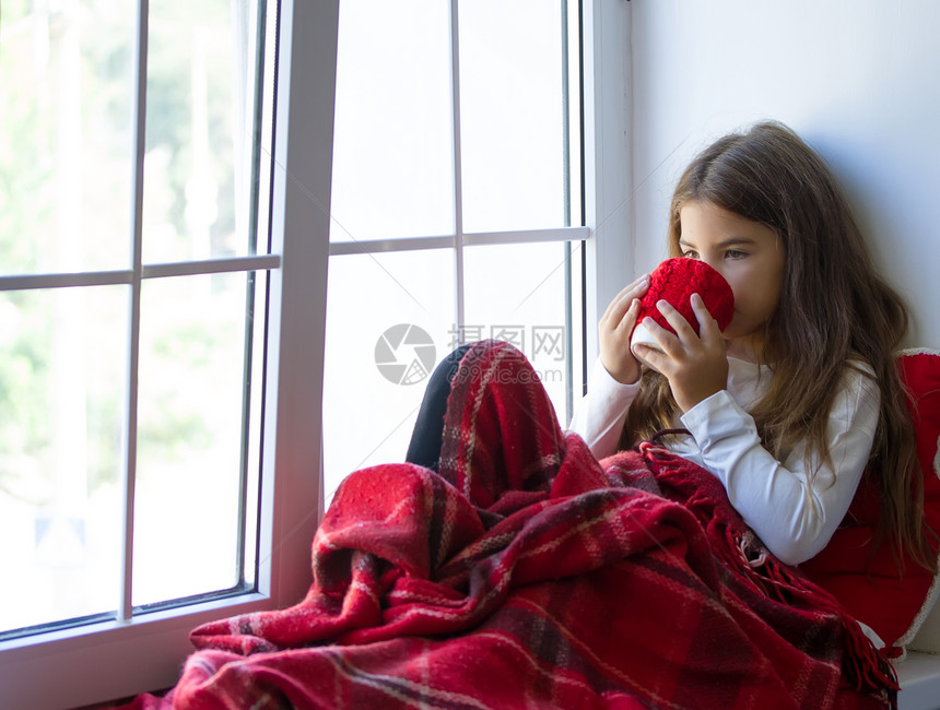 女孩用暖的毯子裹住自己坐在窗边喝杯子上的热饮图片