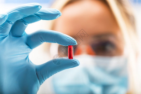 年轻有吸引力的女科学家带着保护眼镜和面罩在制药研究实验室内持有红色透明药片手套上戴指头的红色透明药丸背景图片