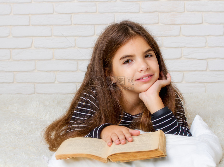 一个小女孩躺在床上拿着一本大旧书在她面前看图片