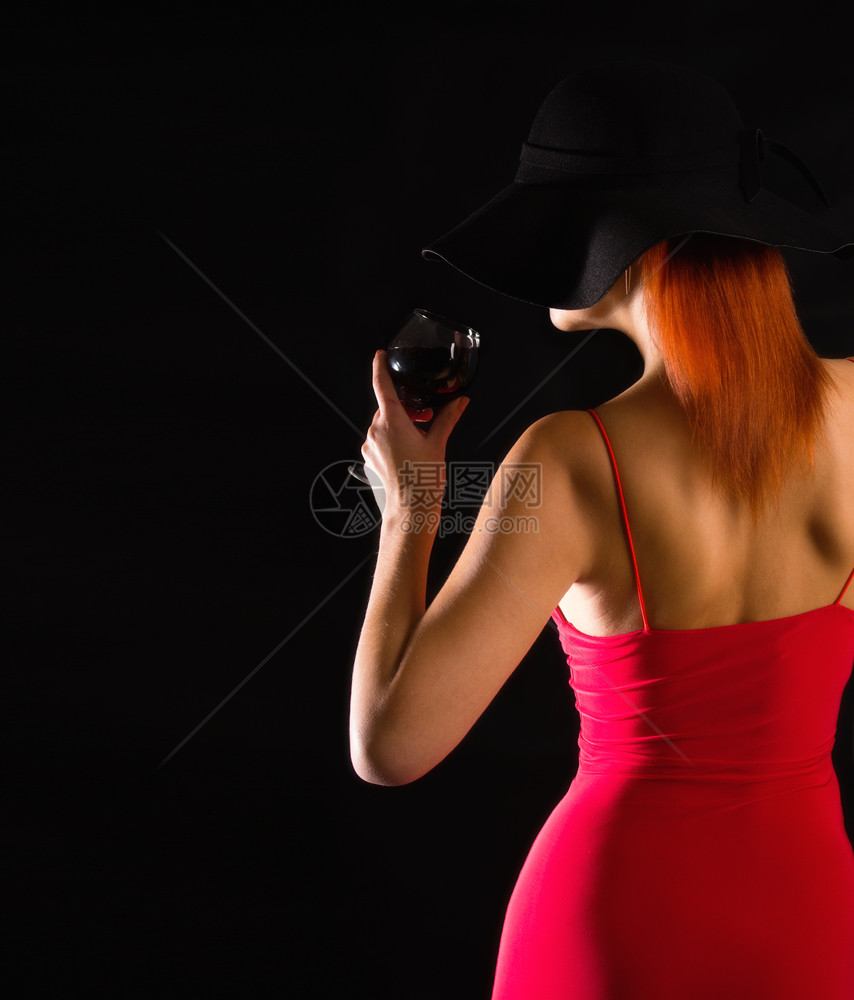 穿着亮红礼服的感女孩穿着黑色宽厚的帽子站在黑暗背景的酒杯上女孩帽子和酒图片
